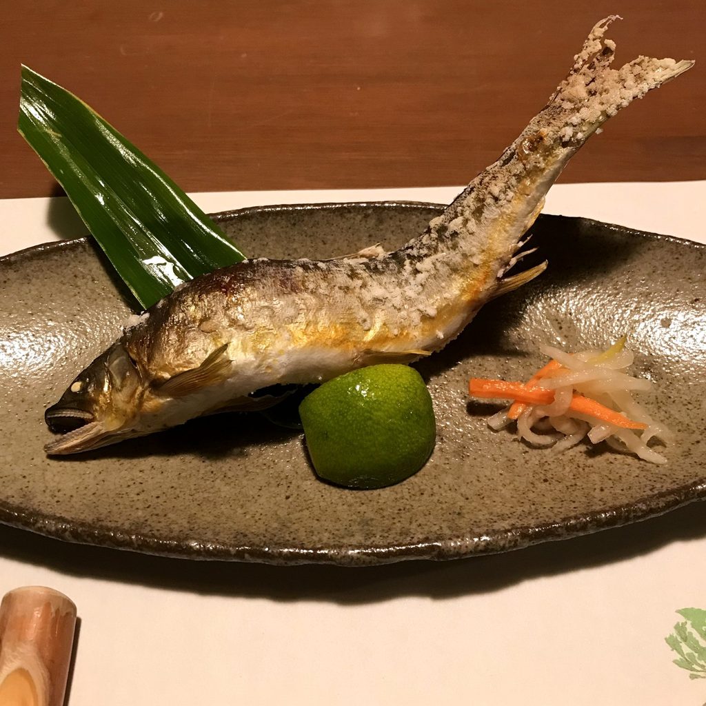 摘み草の宿こまつの夕食の鮎の塩焼き。身をくねらせた形で焼いた鮎に、青い柑橘と酢の物を添えている